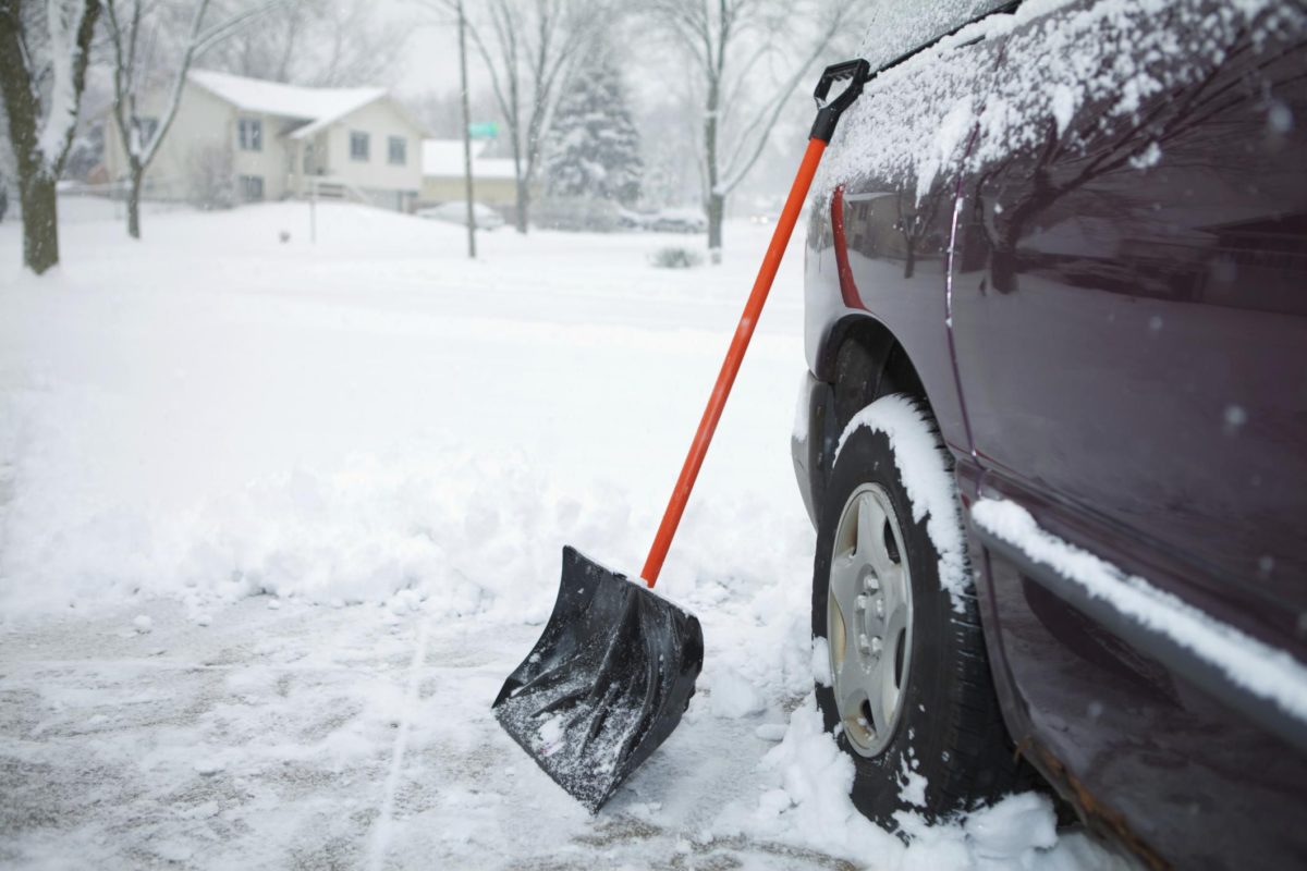 A shovel leans against a car on a snowy driveway